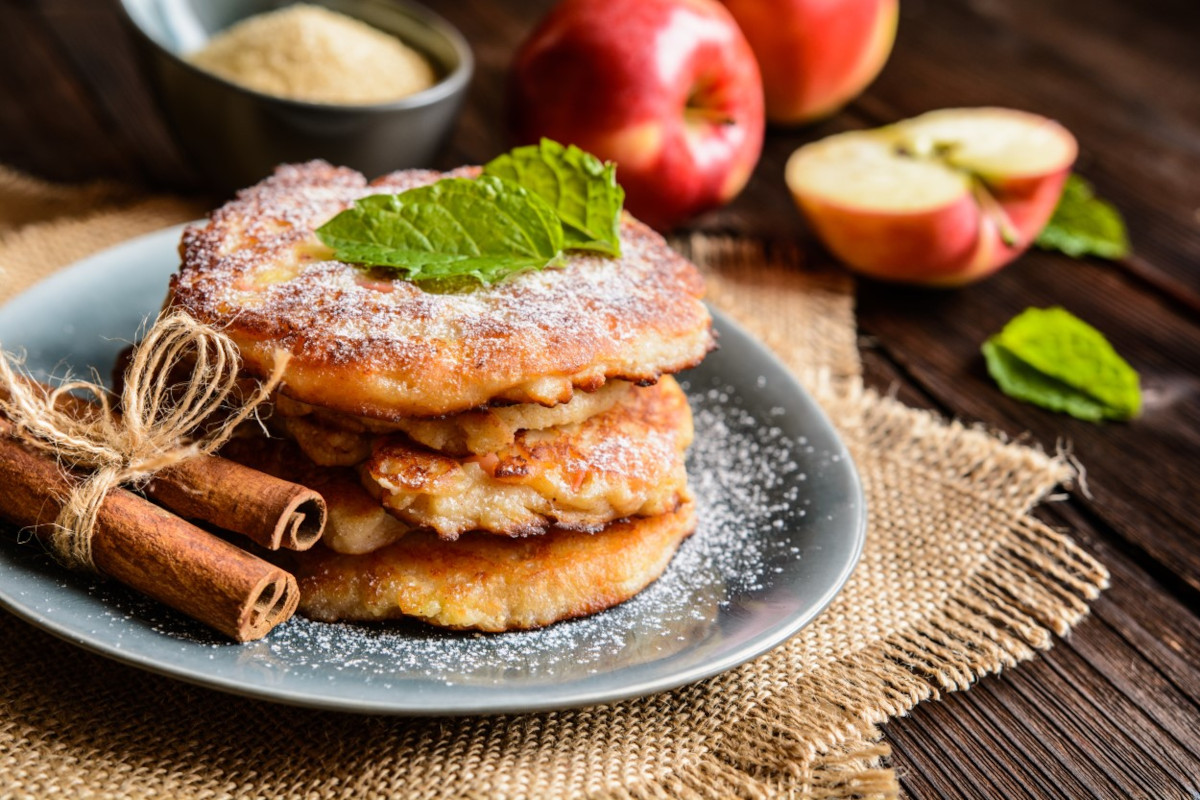 A stack of apple pancakes on a plate next to two cinnamon sticks
