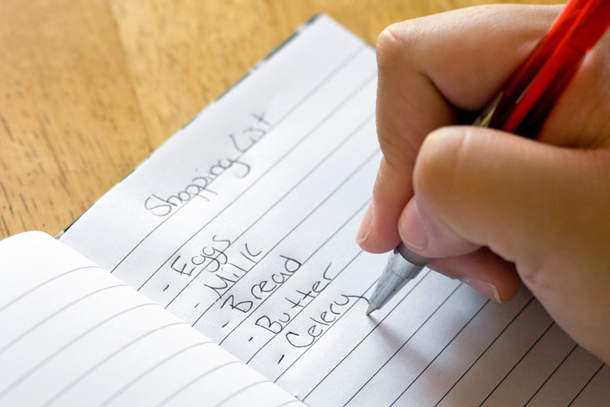A person writing a shopping list that includes eggs, milk, bread, butter and celery