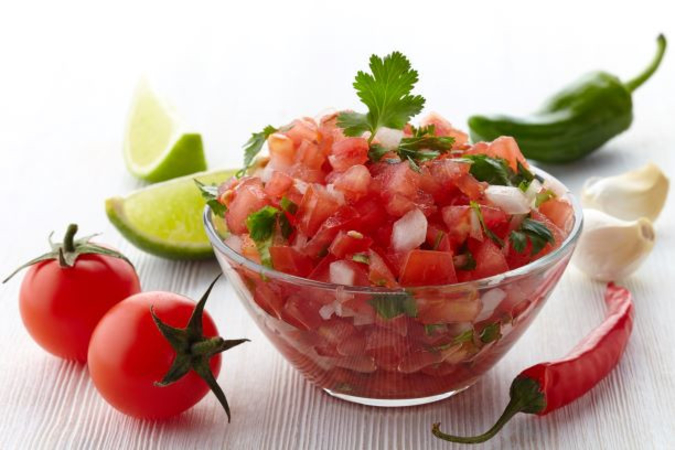 A bowl of salsa with fresh chopped tomatoes, white onion and cilantro surrounded by two whole cherry tomatoes, lime wedges, a chili pepper, two cloves of garlic and a jalapeno pepper
